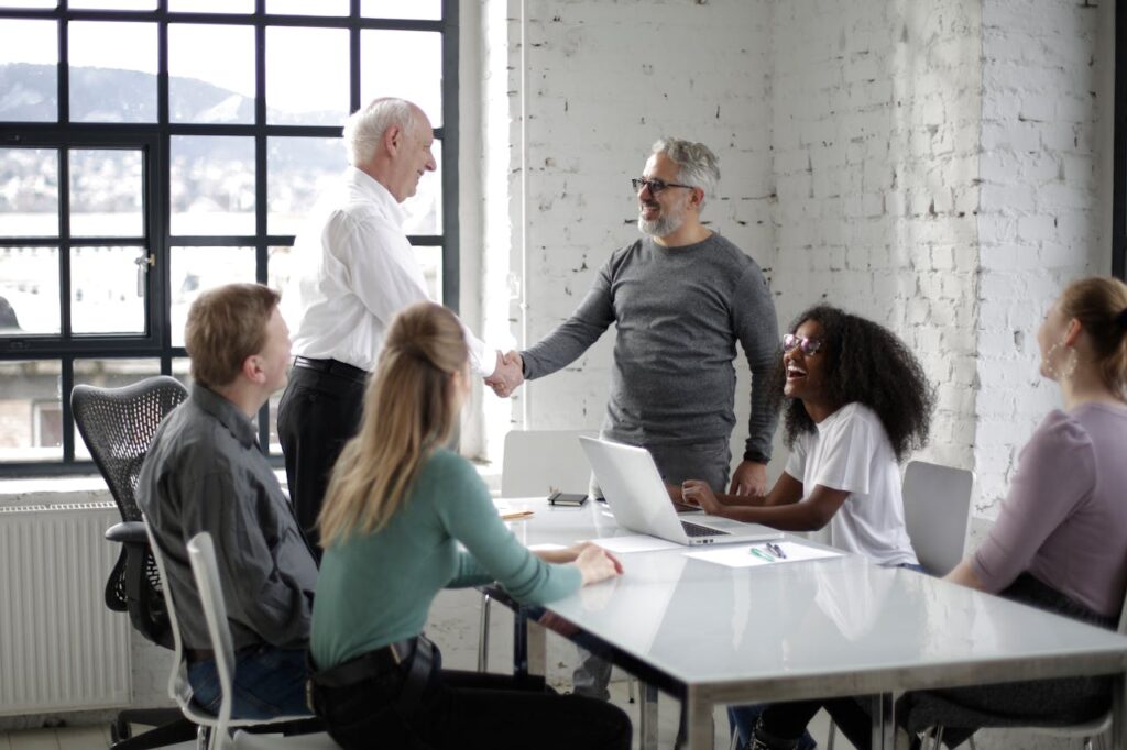 Cheerful male colleagues shaking hands while discussing business ideas with group of multiethnic coworkers gathering around table with gadgets and documents in modern light workspace